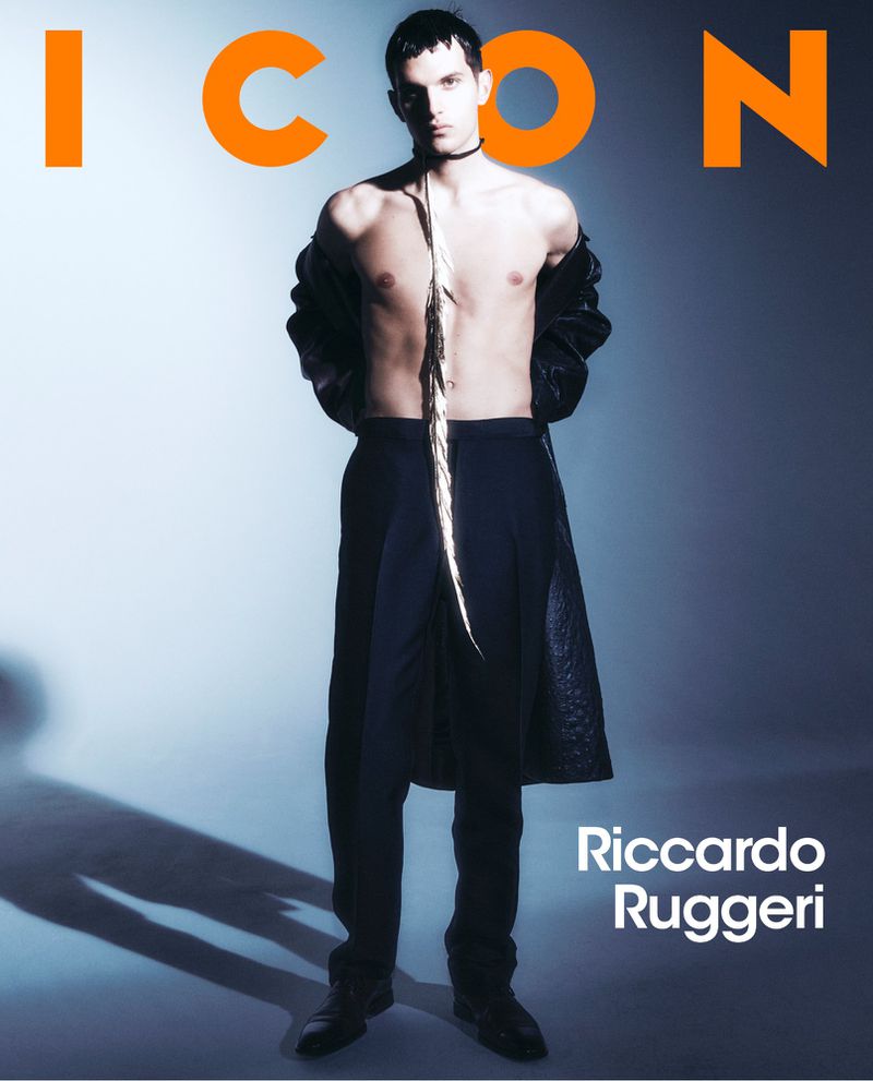 Fashion editorial shot of a topless male model wearing black pants and coat, standing in a photo studio setting with 'ICON' text overlay.