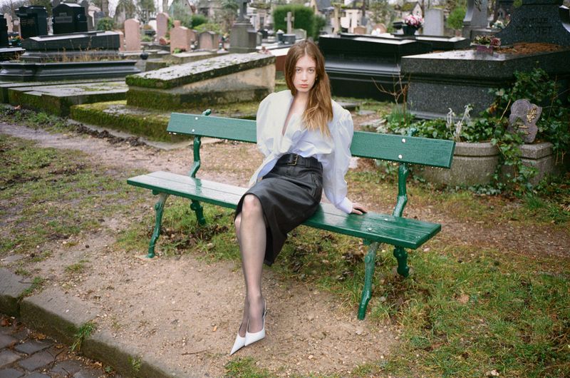 An adult female sits on a green park bench in a cemetery wearing a white blouse, dark skirt, stockings and white heels in daylight.