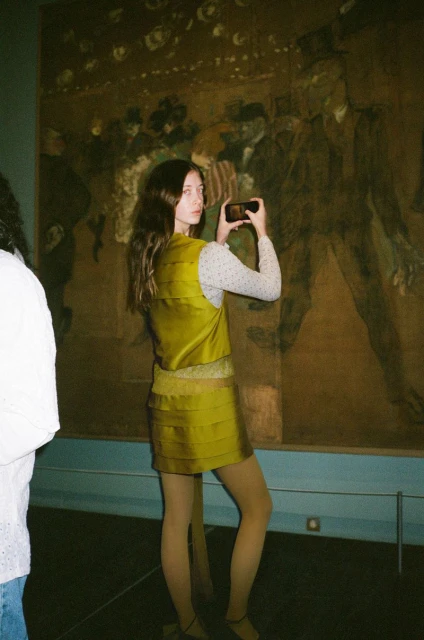 A young woman holds her phone to take a photo of a painting in a museum, wearing a gold skirt and vest and white patterned top.