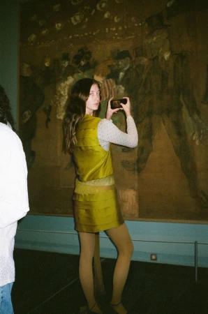 A young woman holds her phone to take a photo of a painting in a museum, wearing a gold skirt and vest and white patterned top.