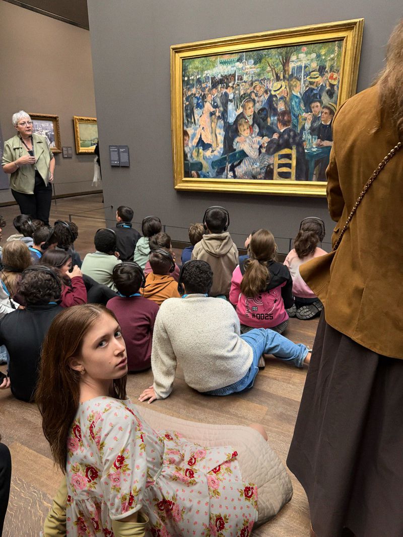 Art history tour at a museum: teacher lectures to diverse group of children sitting on the floor in front of an impressionist painting.