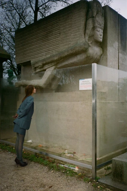 A woman in a suit stands before a weathered stone sculpture of a winged figure, encased in glass, with trees in the blurred background.