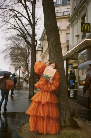Woman in orange tiered dress poses in Paris leaning against tree, holding McDonald's bag. Arc de Triomphe visible in the background.