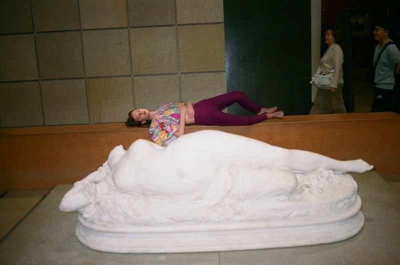 An adult woman poses next to a marble statue in an indoor museum setting with other visitors in the background.