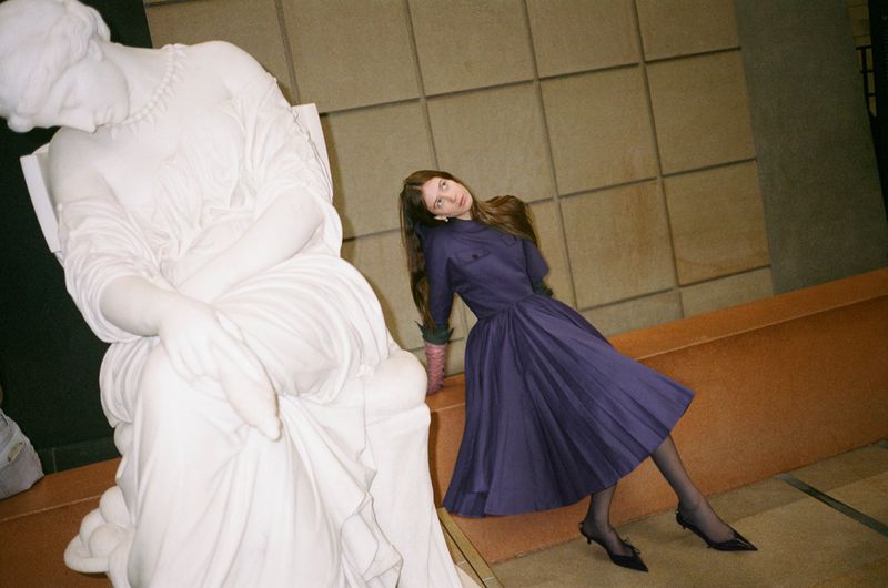 A woman in a purple dress poses next to a white marble statue in an indoor setting with a neutral-toned tile background and warm lighting.