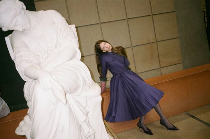 A woman in a purple dress poses next to a white marble statue in an indoor setting with a neutral-toned tile background and warm lighting.