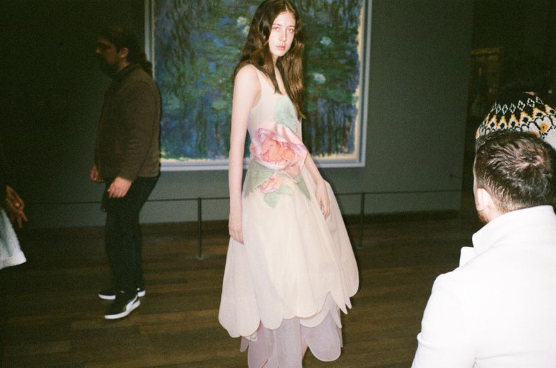 Woman in floral dress at an art gallery, posing near impressionist painting, surrounded by other people admiring artwork.