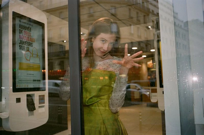 Young woman poses inside a shop window with an advert for handbags. Cars and buildings are visible in soft focus through the glass.
