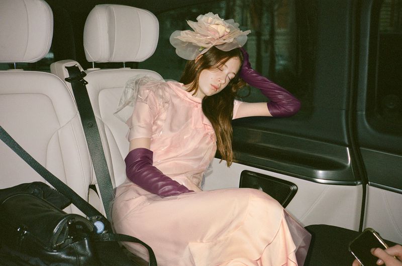 A woman with long brown hair wearing a pink dress and purple gloves sits in a car with a black bag next to her.