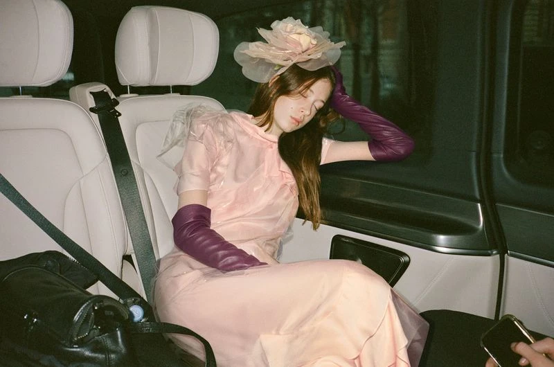 A woman with long brown hair wearing a pink dress and purple gloves sits in a car with a black bag next to her.