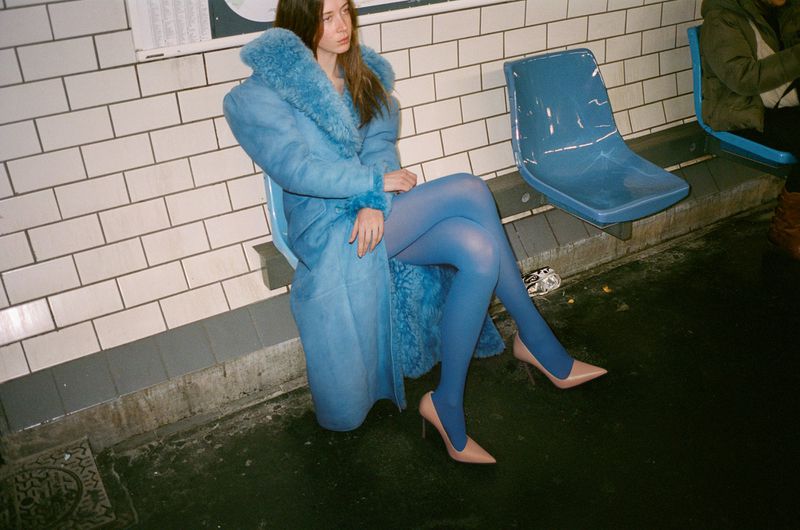 Young woman in a blue fur-collared coat and blue tights sitting on a blue plastic chair in a subway station with tiled walls.