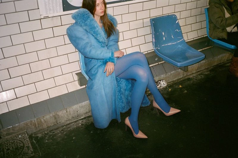 Young woman in a blue fur-collared coat and blue tights sitting on a blue plastic chair in a subway station with tiled walls.