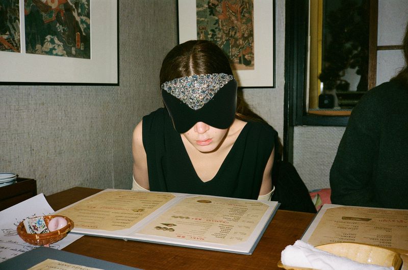 A person with a jeweled blindfold sits at a restaurant table with a menu, creating an intriguing and mysterious atmosphere.