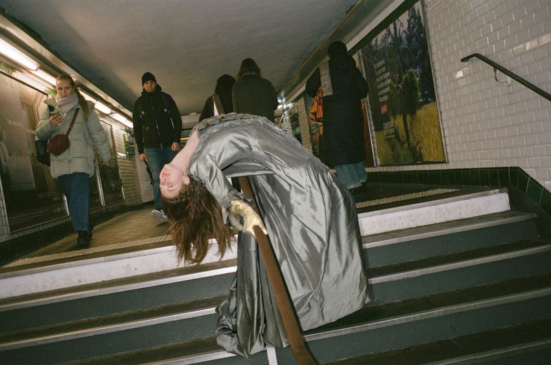 Woman in a long gown leans over a railing in a subway; several people stand in the background in an urban setting. 