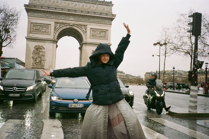 Woman in a black puffer jacket and ballgown skirt joyfully poses in front of the Arc de Triomphe in Paris on a wet, overcast day.