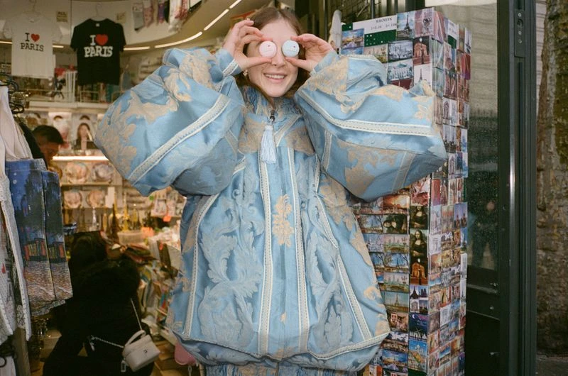 Smiling woman in light-blue patterned coat holds up two round objects at an indoor market with souvenirs and postcards displayed.