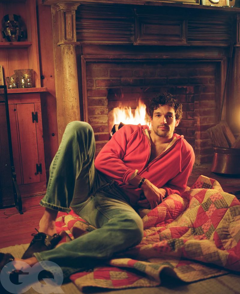 Man in pink hoodie and jeans sits fireside on a quilt. Warm indoor lighting. GQ logo is visible on bottom left corner.  