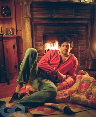 Man in pink hoodie and jeans sits fireside on a quilt. Warm indoor lighting. GQ logo is visible on bottom left corner.  