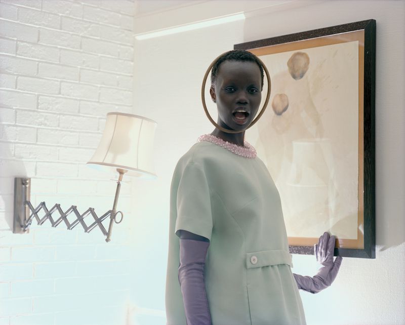 Dark-skinned woman wearing a light-green dress with a beaded collar stands indoors next to a framed painting in a light-filled room.