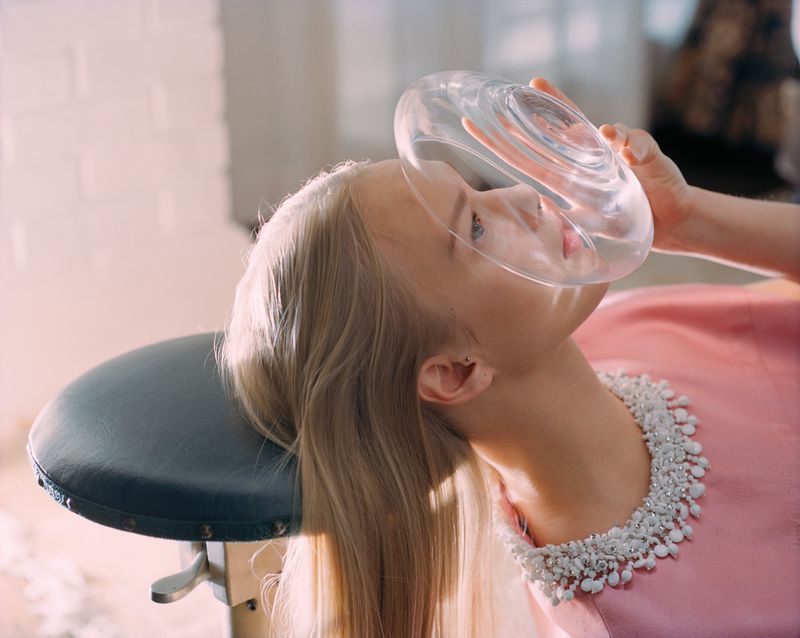 An adult female lays back wearing a pearl necklace and a pink dress, with a clear circular object on her face indoors.