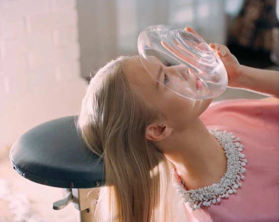 An adult female lays back wearing a pearl necklace and a pink dress, with a clear circular object on her face indoors.