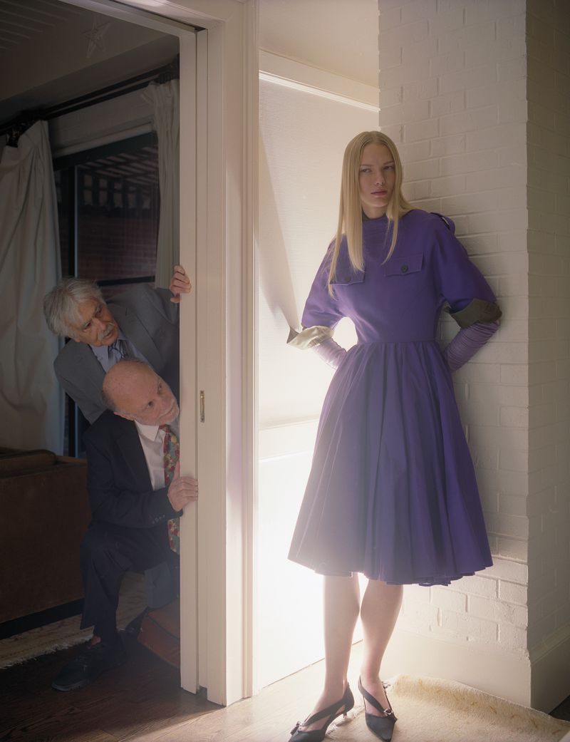 Fashion shot of a woman in a purple dress leaning against a wall, with two men peering out from behind a doorway in the background.