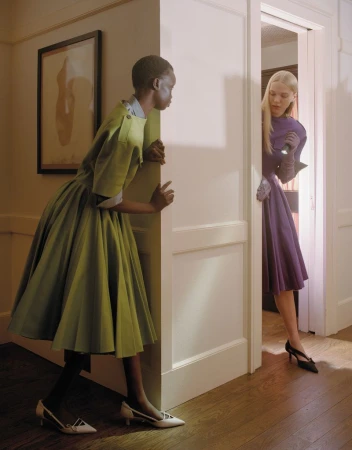 Two fashion models in mid-century dresses and low heels positioned on opposite sides of a white door frame in an indoor setting.