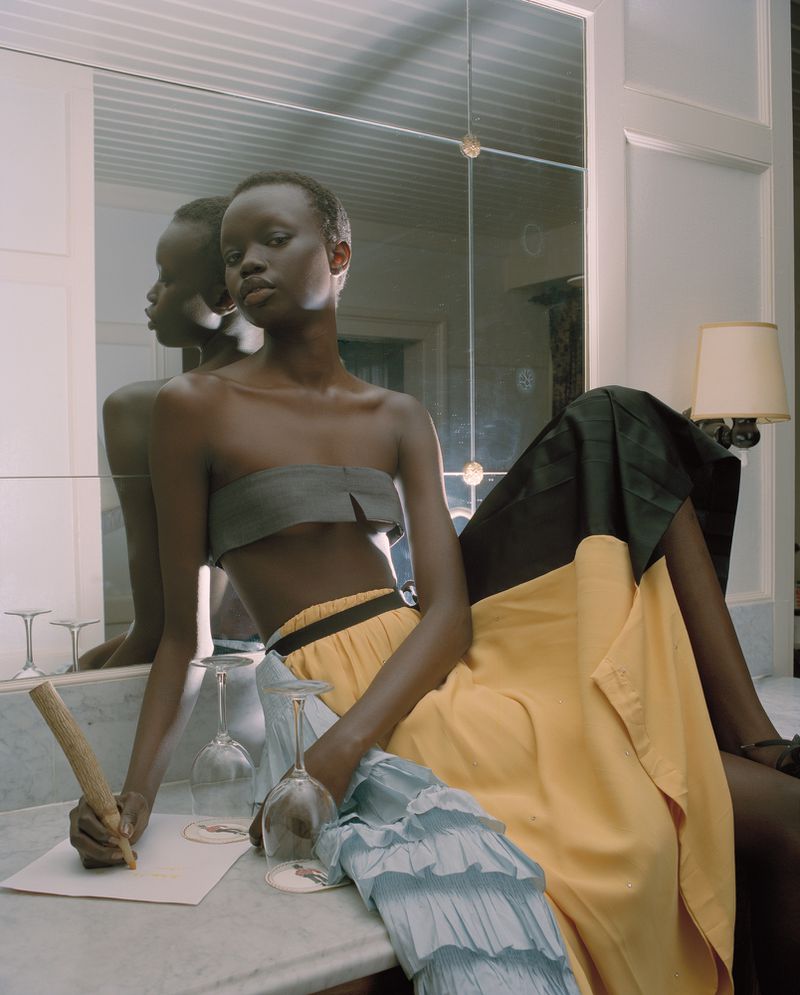 Elegant woman in grey strapless top and yellow skirt seated at marble vanity, signing document with pen in a modern indoor setting.