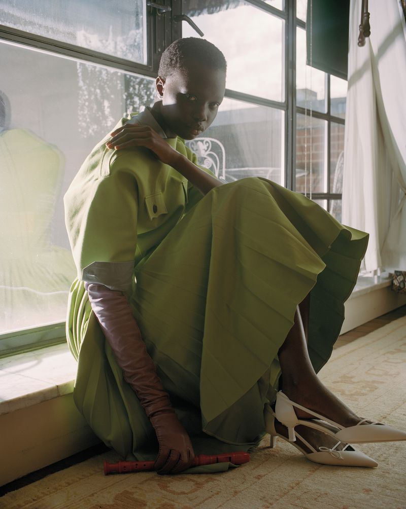 A person wearing a green dress and shoes sits near a window with long gloves, indoors, with a musical instrument.