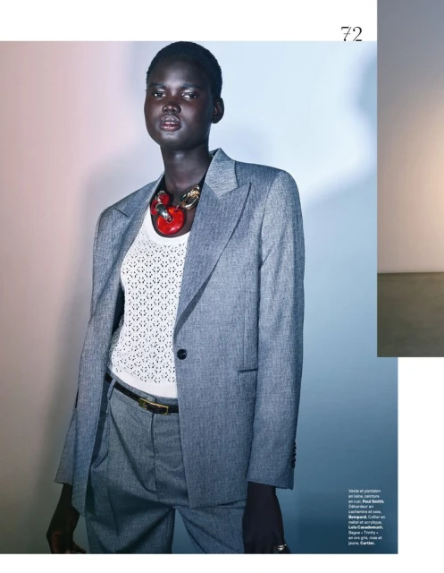 Fashion editorial shot of a Black woman in a grey suit with a large red necklace, posing in a studio against a gradient background.