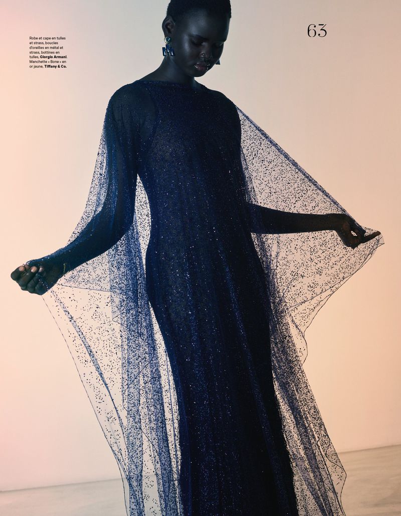 An African-descent adult female fashion model poses in a flowing dark blue gown with cape sleeves in a minimalist, studio-lit setting.