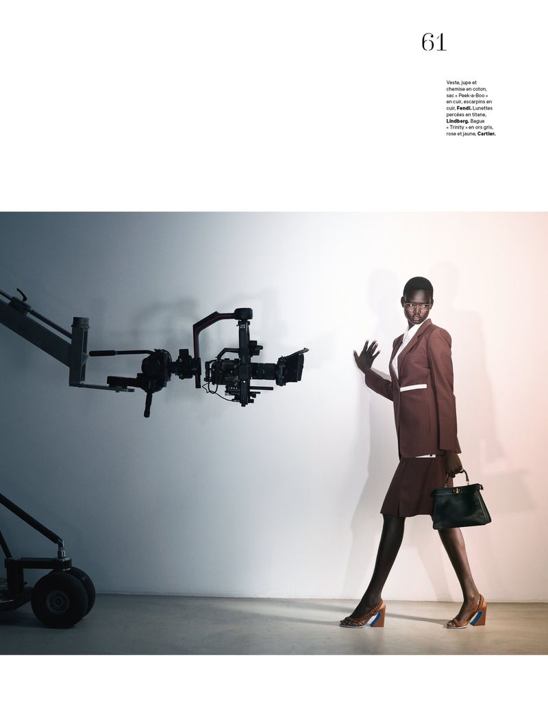 Fashion shot featuring an African woman in a brown skirt suit posing next to professional film camera equipment against a neutral backdrop.