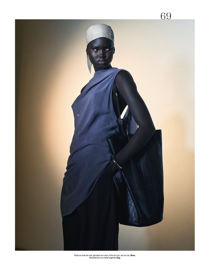 Elegant dark-skinned woman poses in a blue top, black pants, and head wrap, carrying a black leather tote bag in a studio setting.