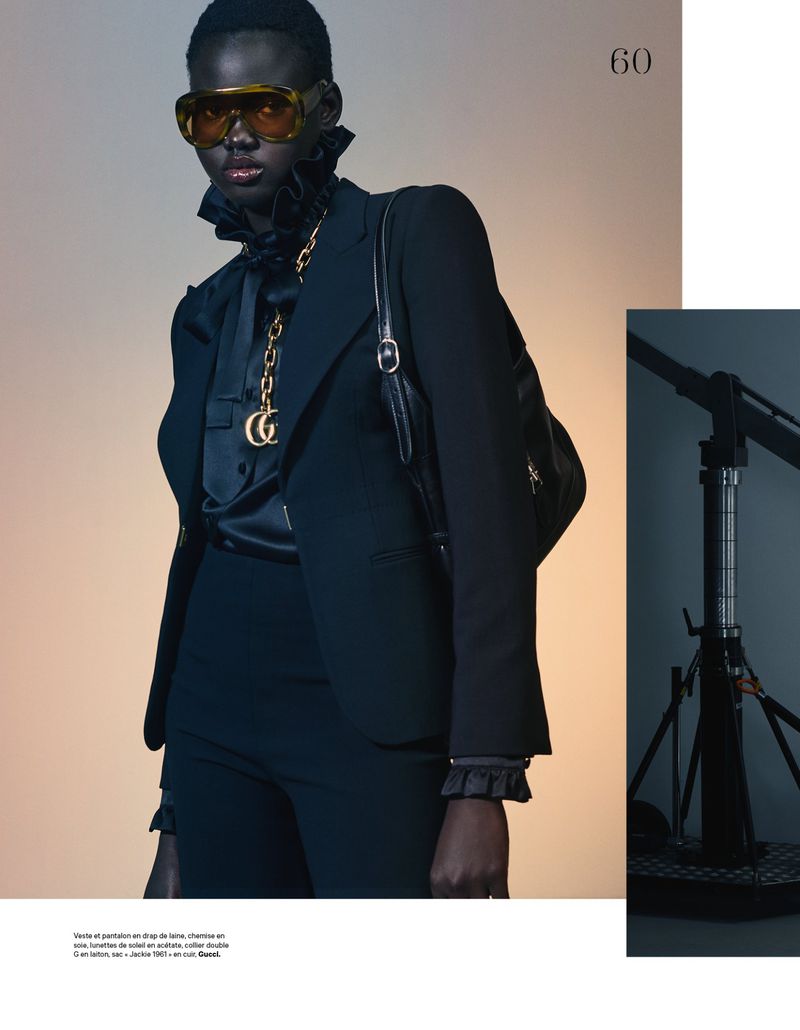 Fashion editorial shot of a woman with dark skin in a black suit, sunglasses, and gold necklace, standing in a photo studio.