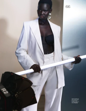 Fashion editorial shot of an African female model in white suit and black bra, accessorized with crystal earrings, and a light stand. 