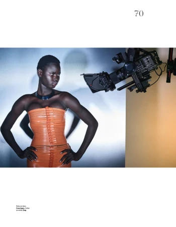 Model Nyakong Chan wearing an orange strapless top and dark choker in a studio setting with a professional camera visible