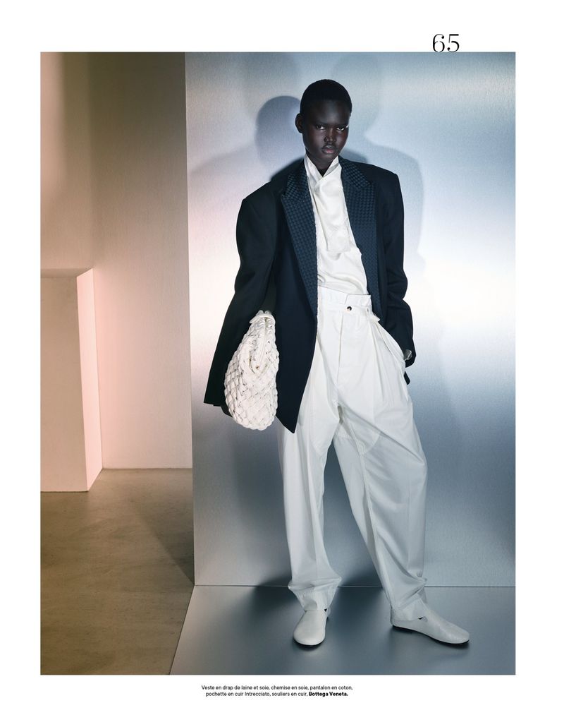 A black model poses in a studio, wearing a black blazer, white shirt, and wide-leg trousers, holding a woven bag, under bright studio lighting.