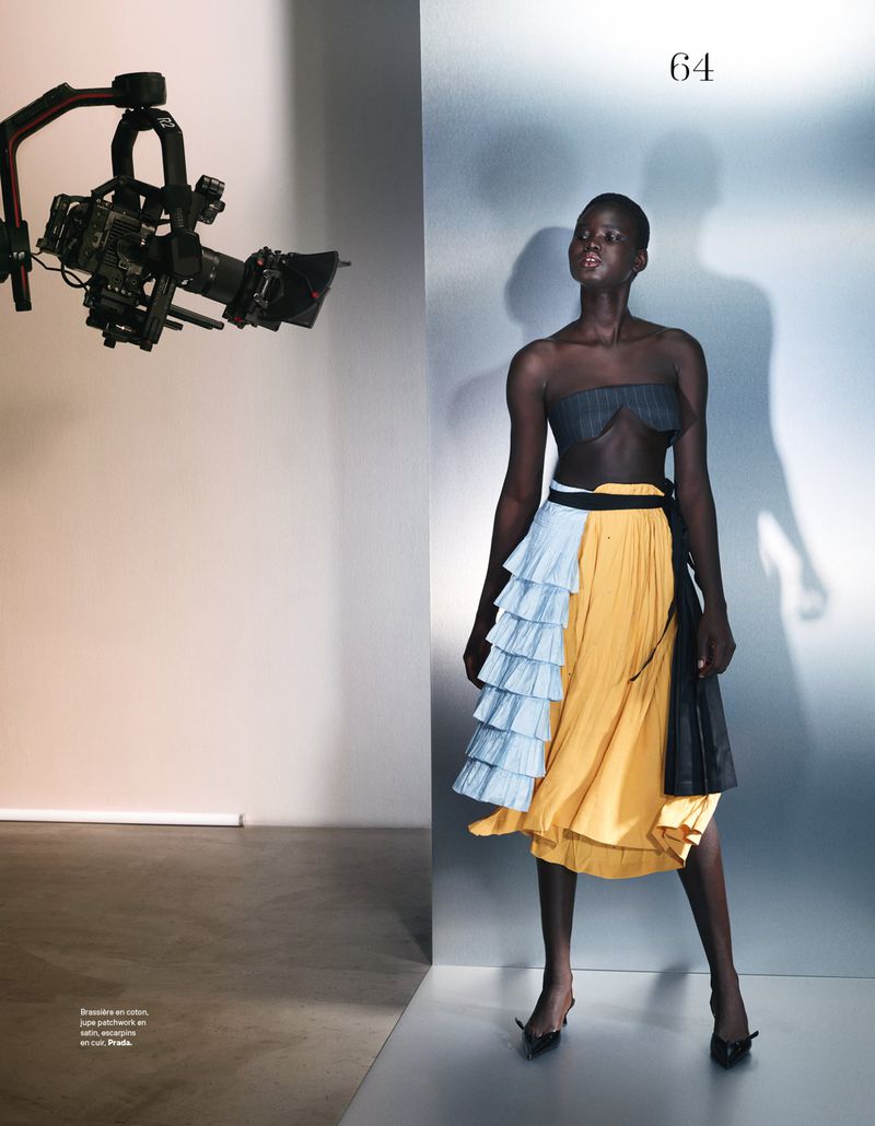 Fashion editorial shot of a Black model wearing a designer dress, standing in a studio setting with a camera rig visible in the background.