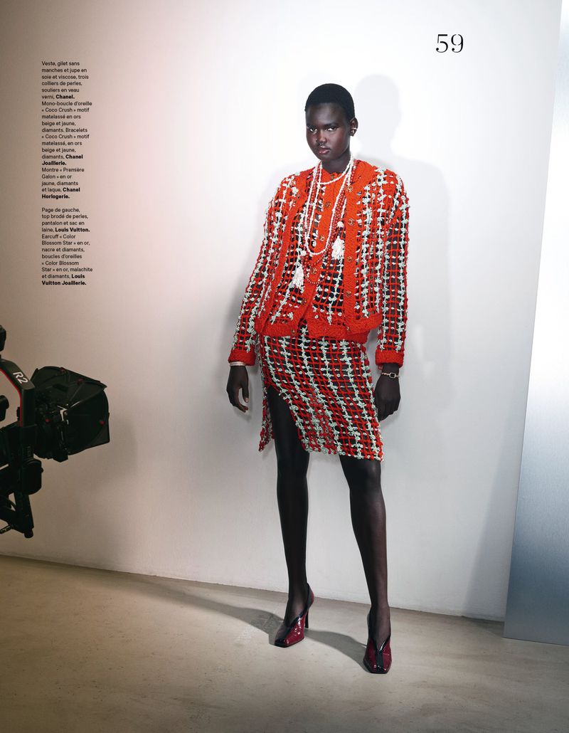 Model standing against a plain white studio wall wearing a red and white patterned skirt suit with layered pearl necklaces.