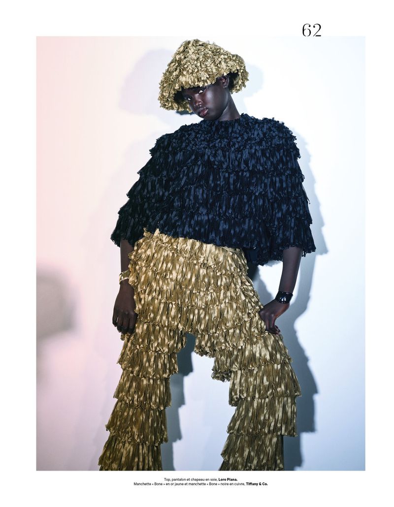 Editorial fashion shot of a Black model wearing a fringed black top and gold tiered fringed pants with a gold hat on a white background.