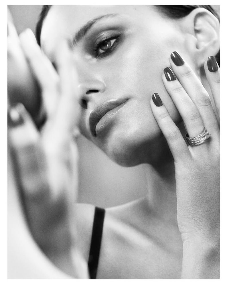 Close-up black and white portrait of a woman with dark red nails, wearing a ring, looking in a mirror with a neutral expression.