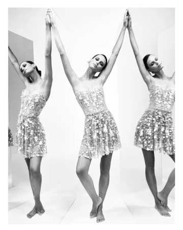 Monochrome fashion shot: a woman in a textured mini-dress with arms raised, reflected in mirrors creating a multiplied effect in a studio setting.