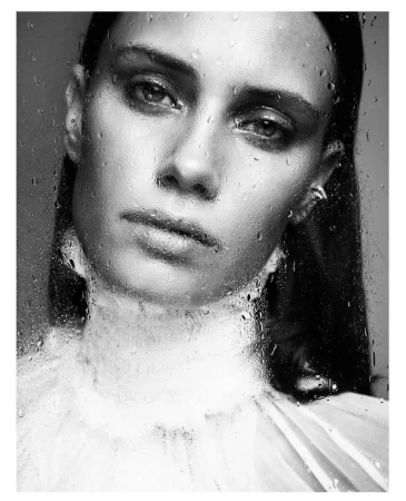 Close-up portrait of a woman viewed through wet glass with water droplets, featuring soft monochrome tones.