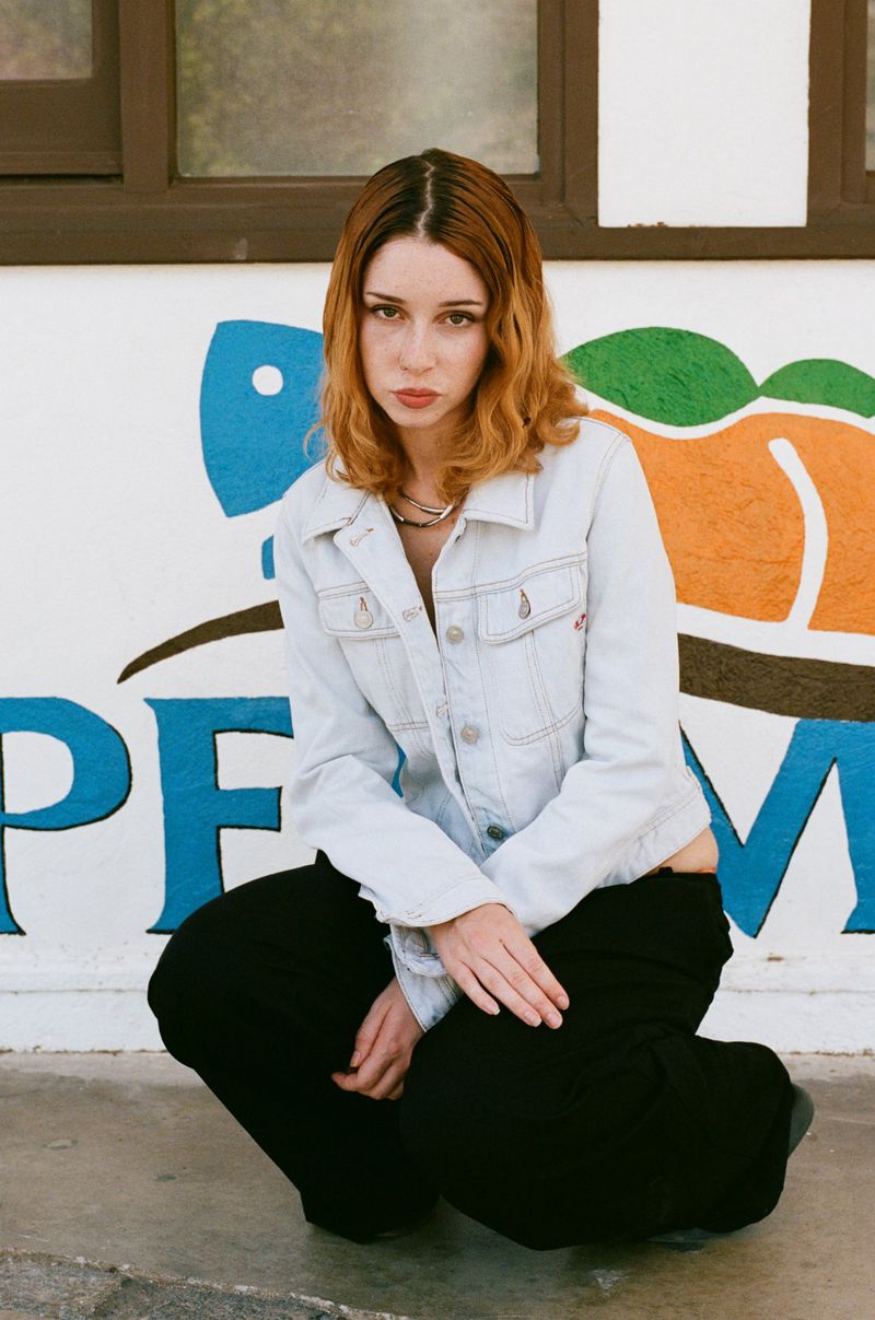 A young woman with auburn hair and a light denim jacket is squatting in front of a graffiti-covered wall in an outdoor setting.