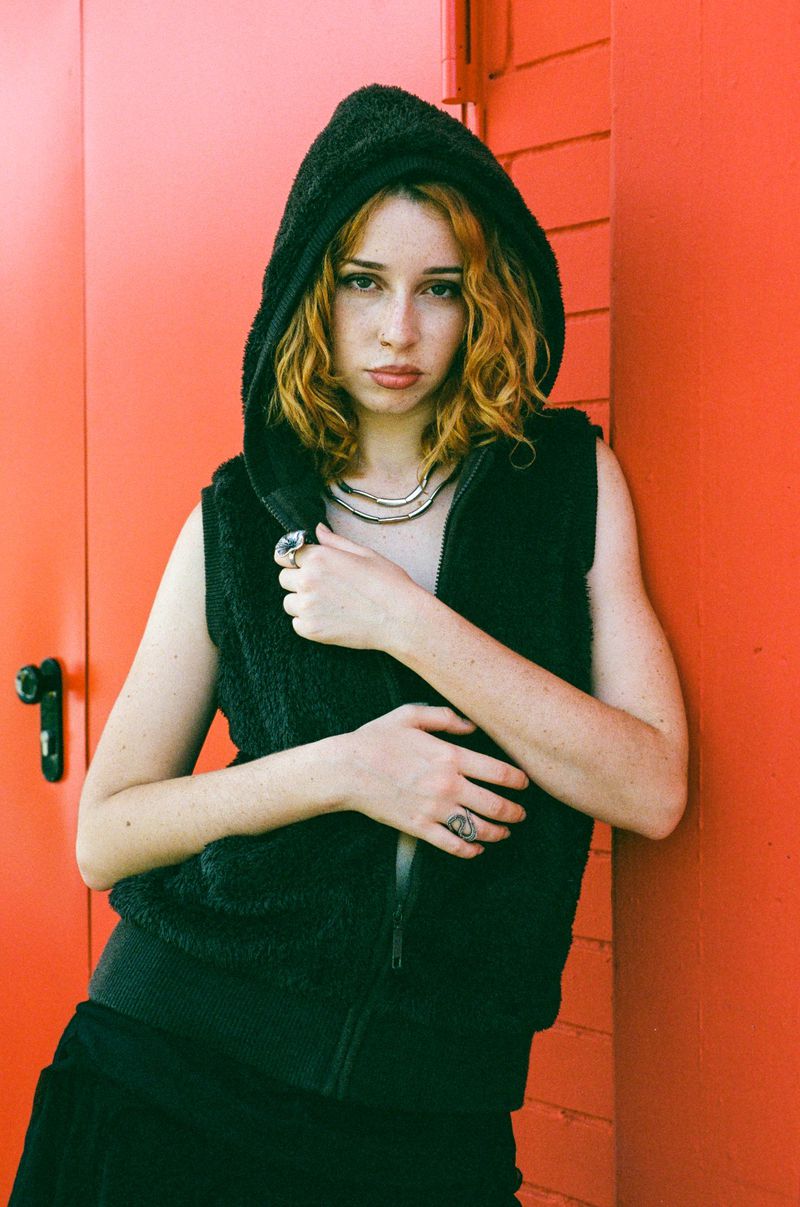 Young woman with freckles wearing a dark hooded vest poses against a bright orange exterior wall with a metal handle.