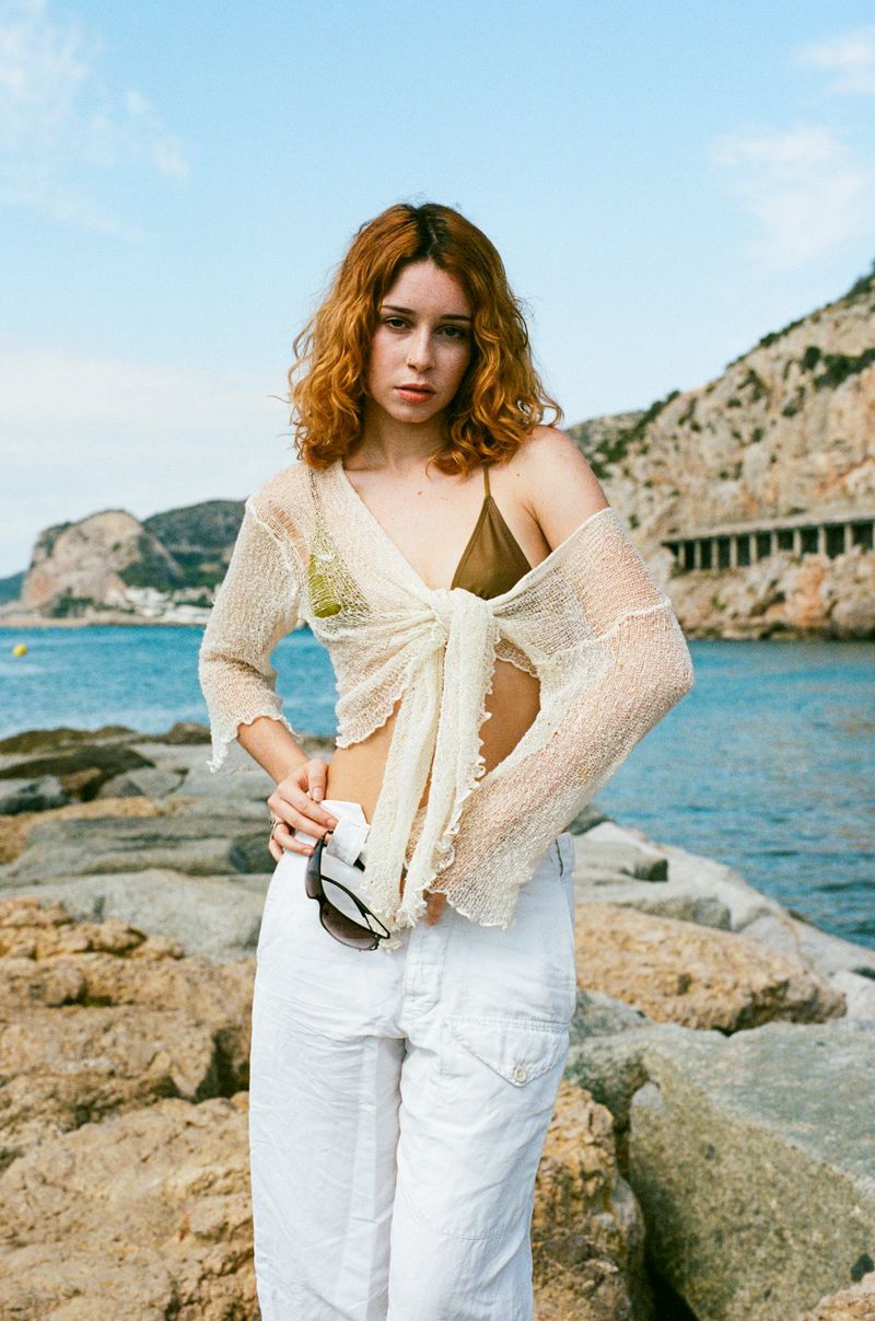 Young woman in casual beach attire poses on a stone pier with mountains and blue sky in the background, creating a serene coastal portrait.