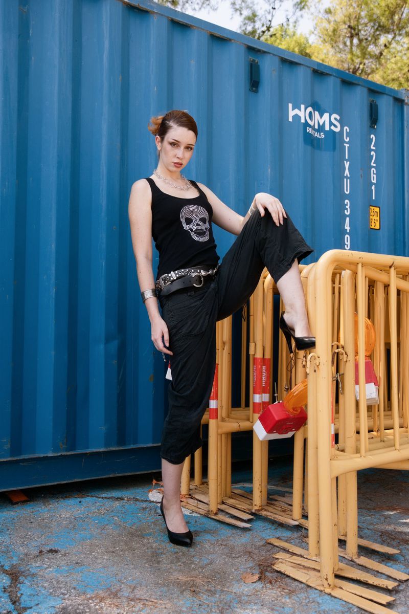 Adult woman poses in black top with skull print, culottes, and heels against a blue shipping container and yellow metal barriers.