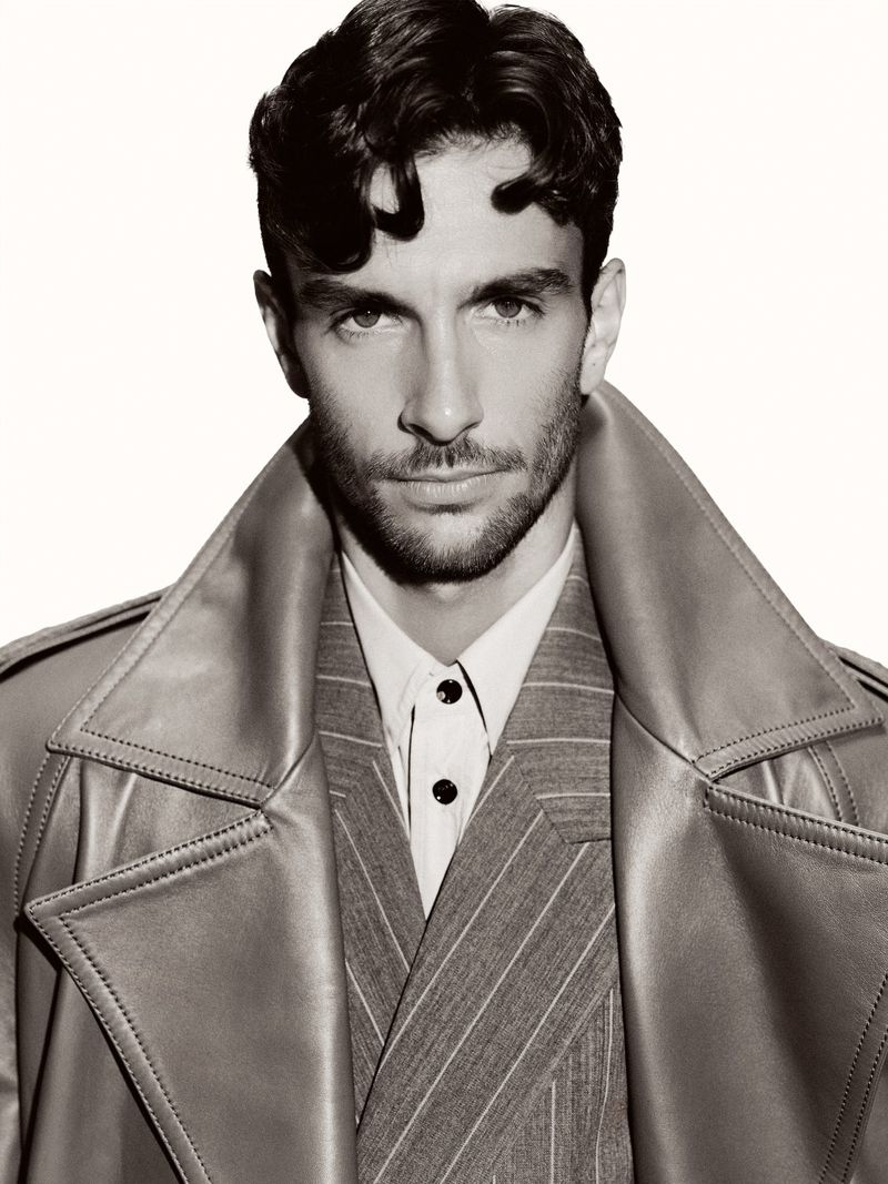 Portrait of an adult male model with wavy hair wearing a trench coat and pinstripe suit against a clean, light background.