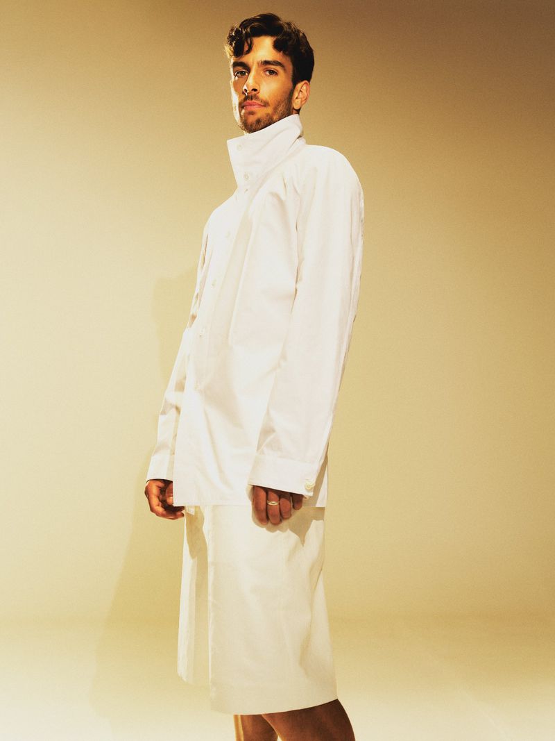 Portrait of a young adult male in a white outfit against a neutral gradient background in a studio setting. Fashion shot.