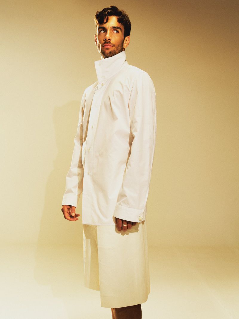 Young man stands in a photo studio wearing an oversized white shirt and matching shorts, looking off to the side with a neutral expression.
